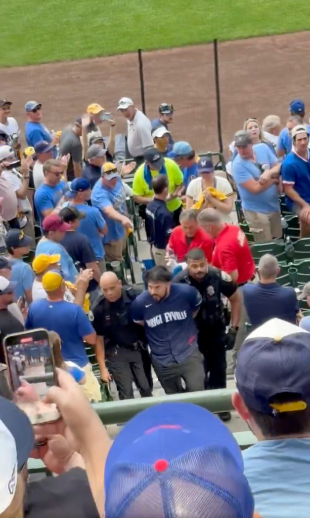 A man wearing a "WRIGLEYVILLE" shirt being escorted by security at a baseball game.